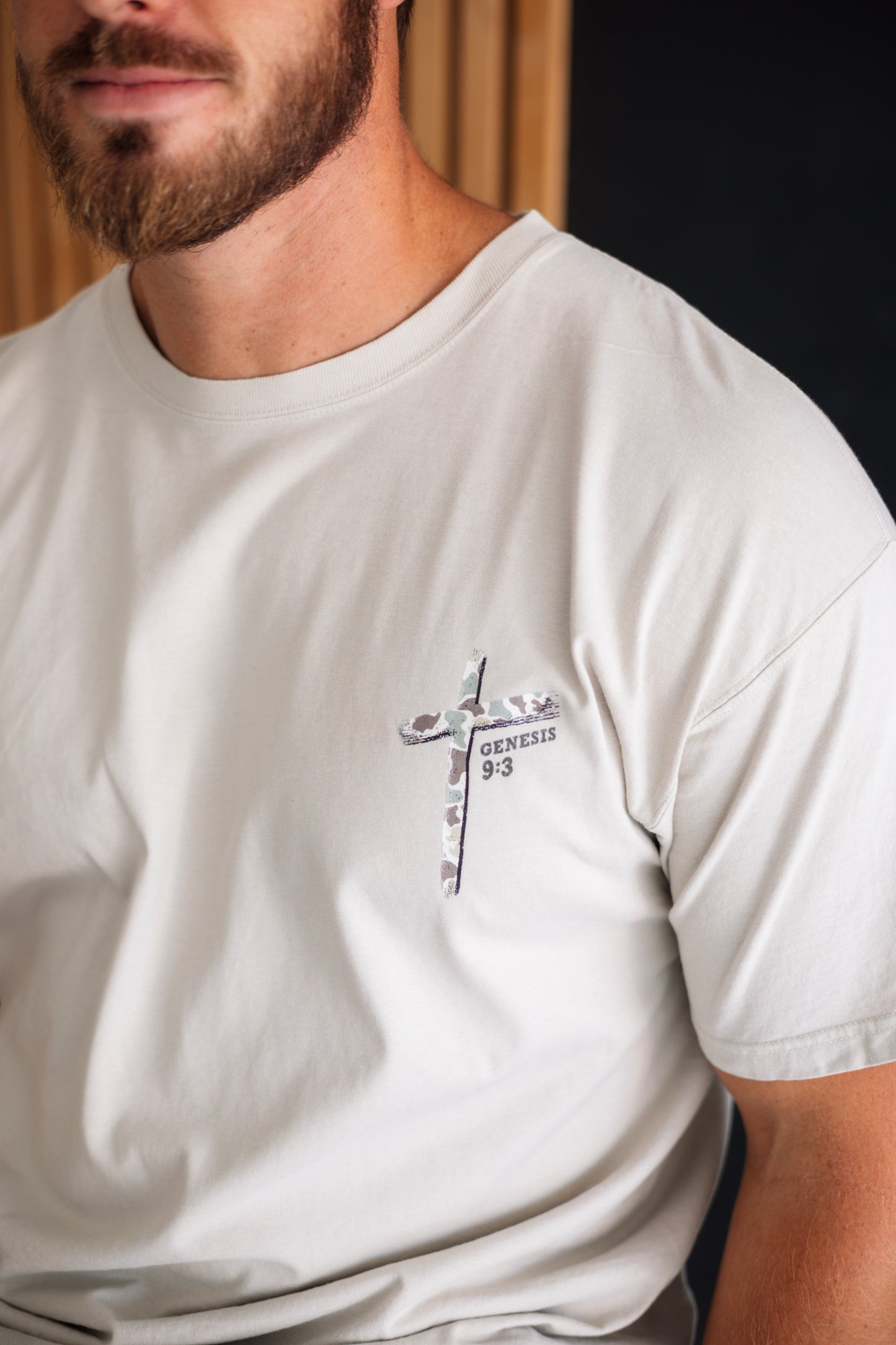A close-up of a man wearing a light beige crew-neck t-shirt with a small cross graphic on the left chest containing the text 'GENESIS 9:3'.