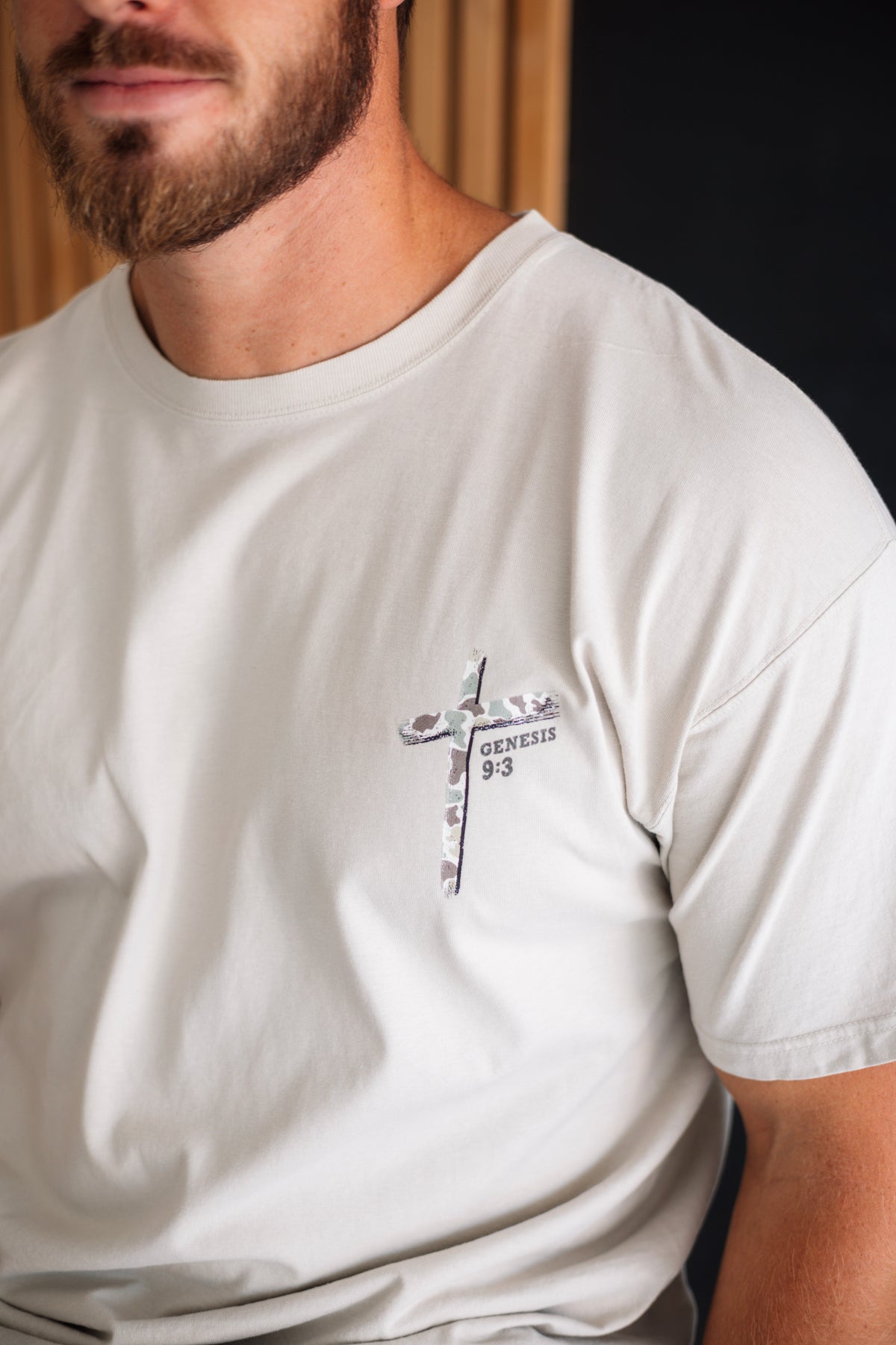 A close-up of a man wearing a light beige crew-neck t-shirt with a small cross graphic on the left chest containing the text 'GENESIS 9:3'.