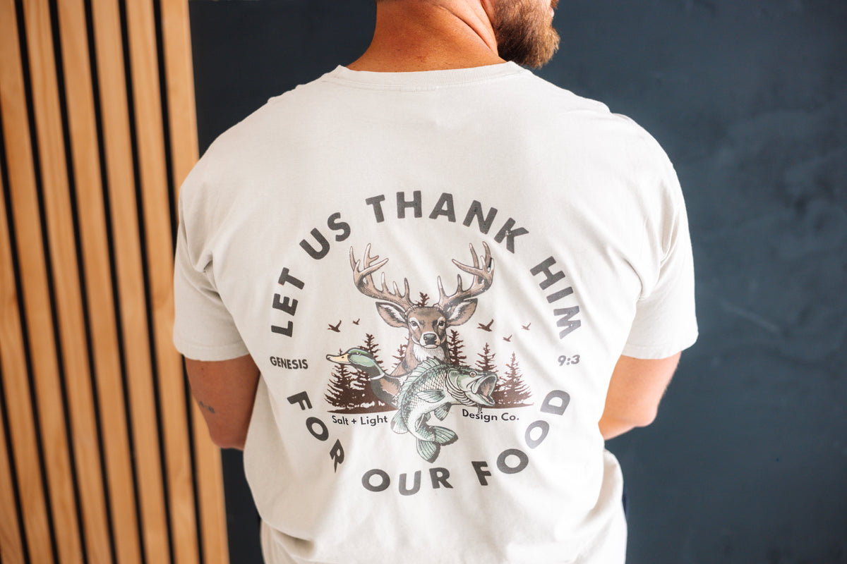 Back of a cream Christian T-shirt featuring “Let Us Thank Him for Our Food” Genesis 9:3 with deer, duck, fish, and outdoor graphics.
