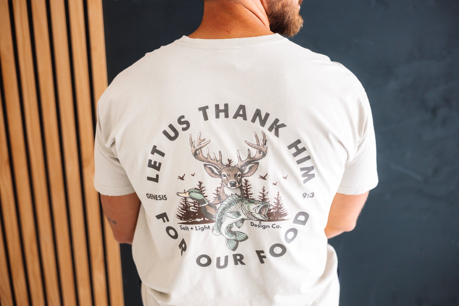 Back of a cream Christian T-shirt featuring “Let Us Thank Him for Our Food” Genesis 9:3 with deer, duck, fish, and outdoor graphics.